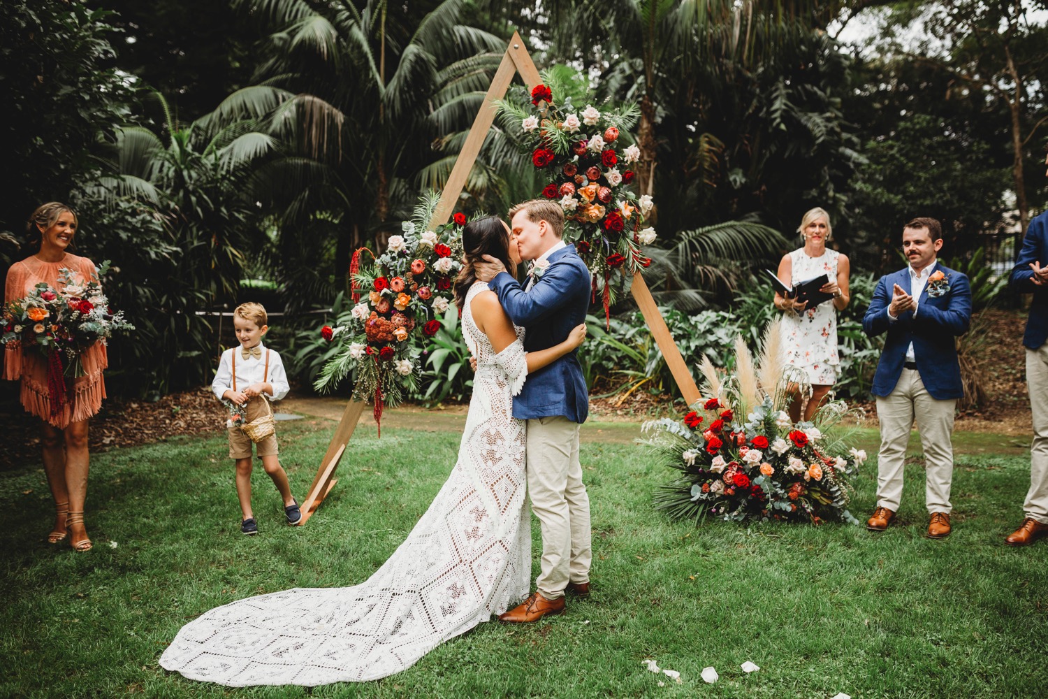 Newly married couple embracing in front of floral arch in the gardens. The bride wears a white dress and the groom wears a blue blazer and light pants. The celebrant wearing a light coloured dress is the background, as well as a bridesmaid in a light colour dress, a groomsman in an outfit similar to the groom and there is a pageboy wearing a white shire and brown shorts. Everyone is very happy.