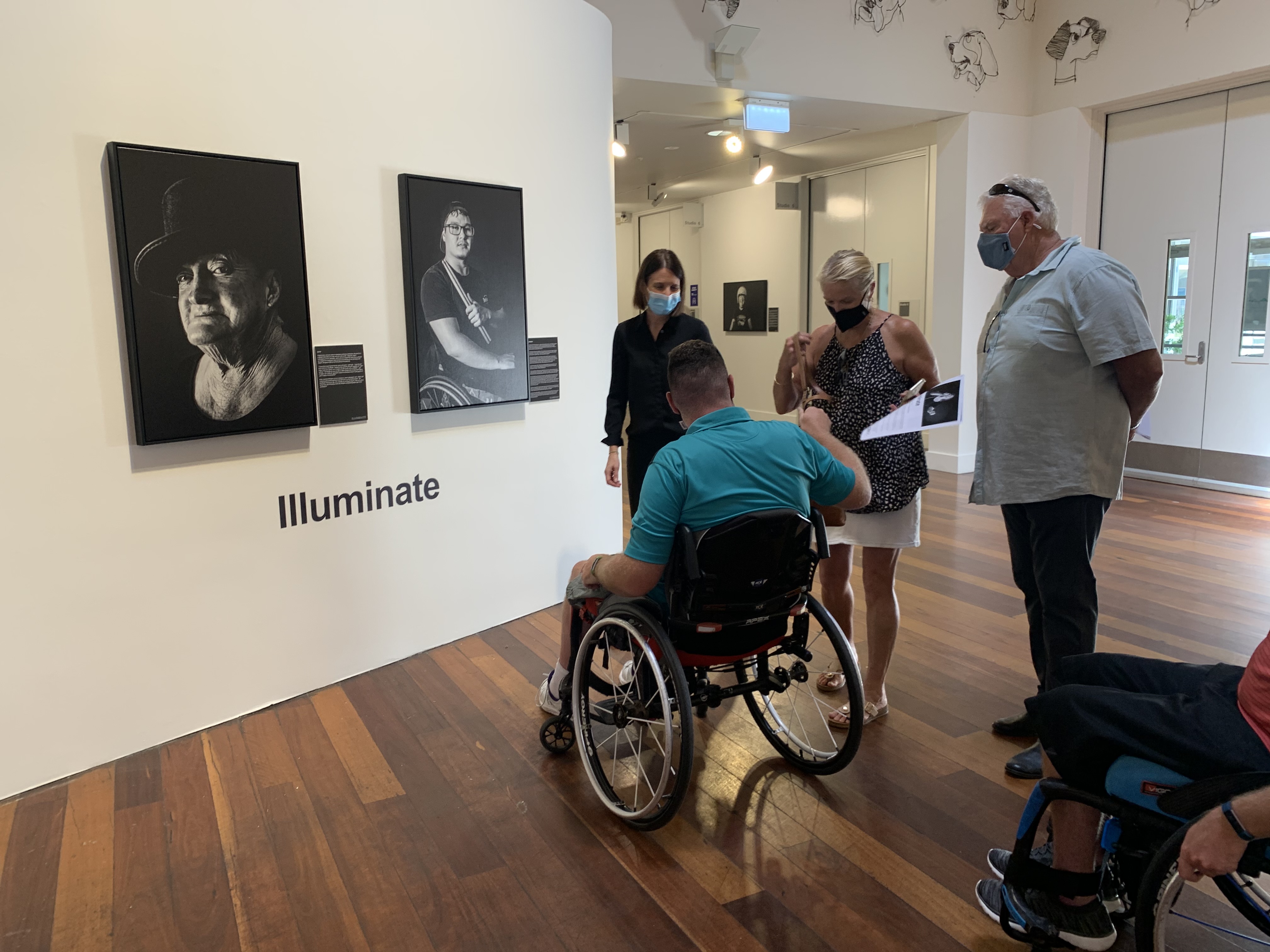 Art gallery displaying black and white portraits with several visitors observing. Two people are in wheelchairs. 