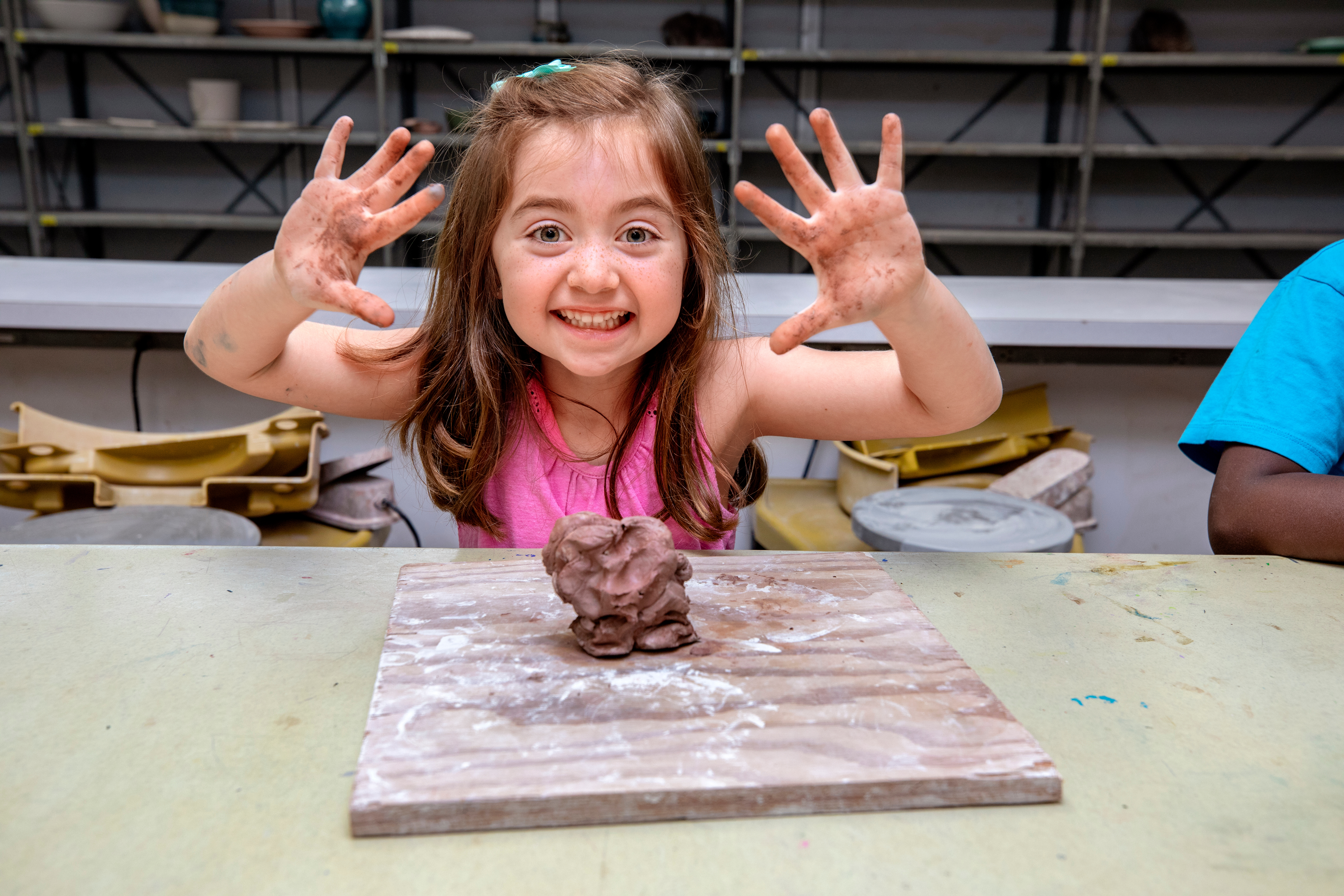 Girl child in clay class, smiling at camera with hands open.