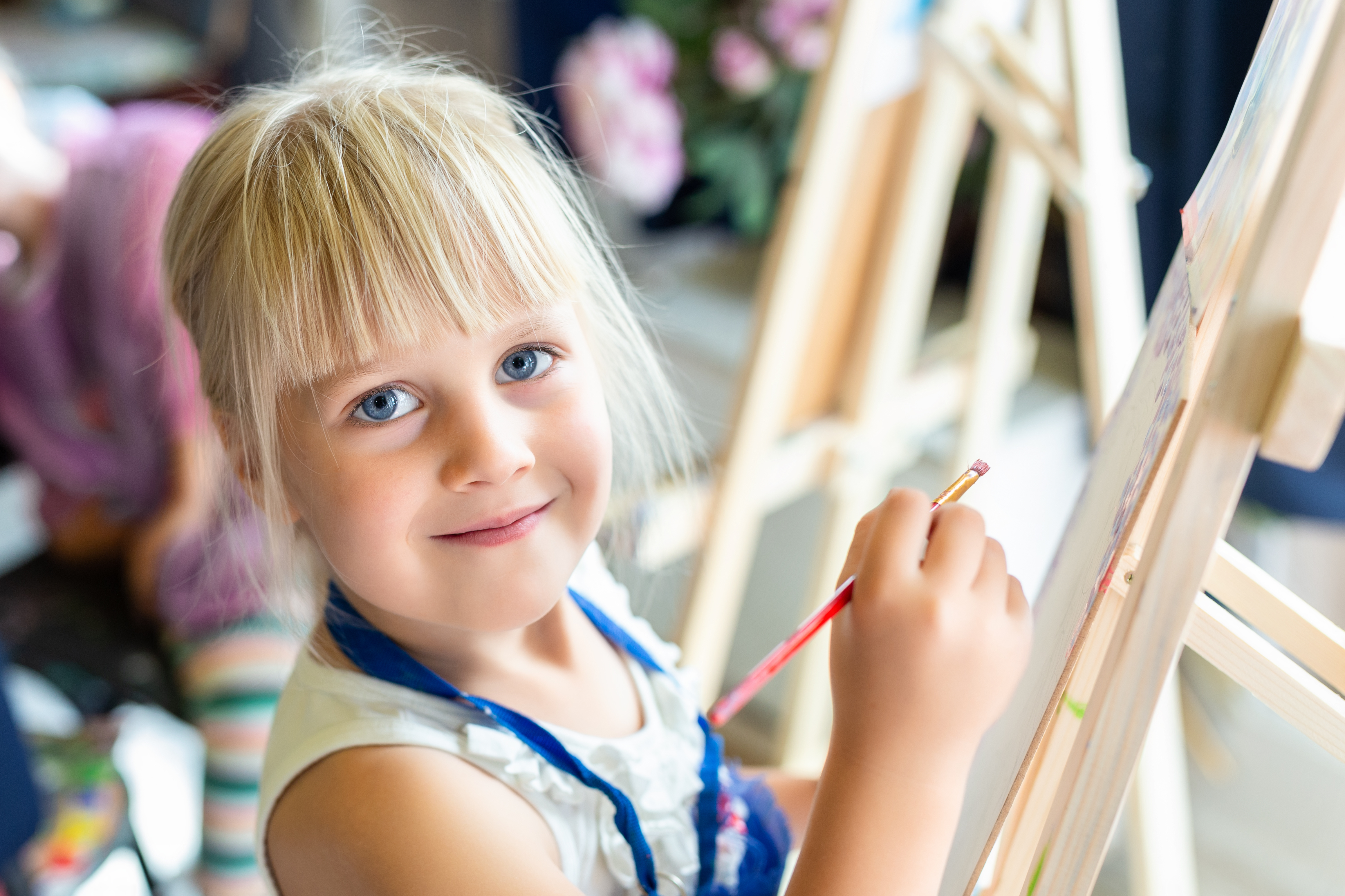 Blonde female child standing at painting easel smiling at camera.