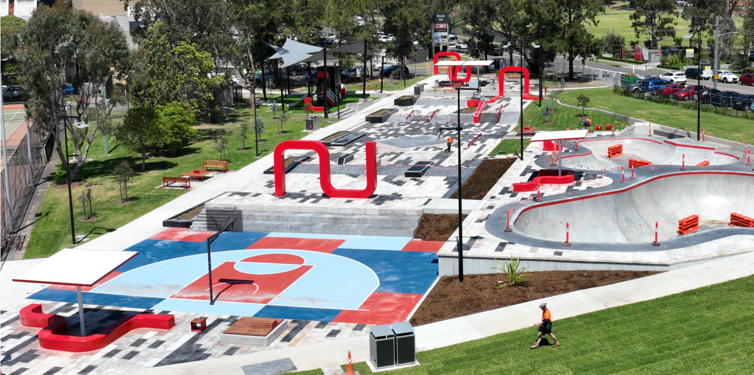 aerial view of plaza with half basket ball court in foreground, skate bowls on right side and red sculptural elements in green parkland