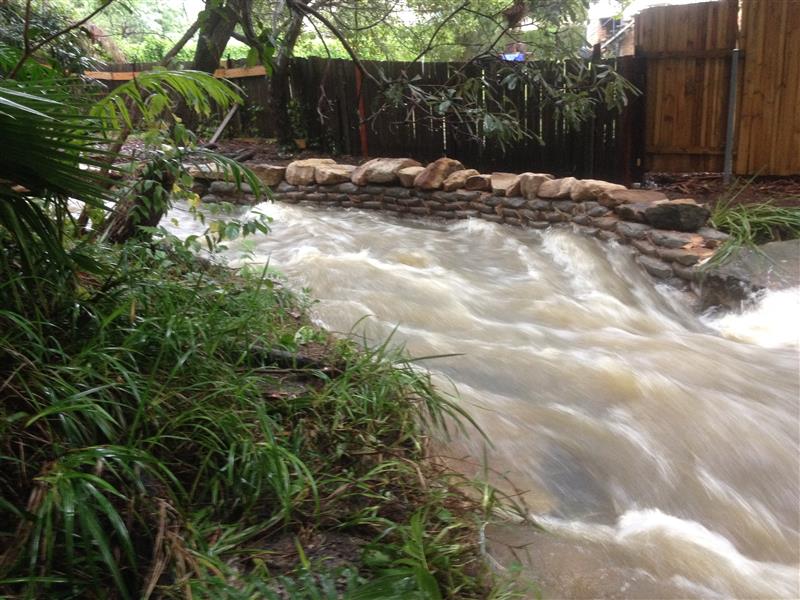 Rushing water, rocks and garden beds surrounding, residential fencing in background