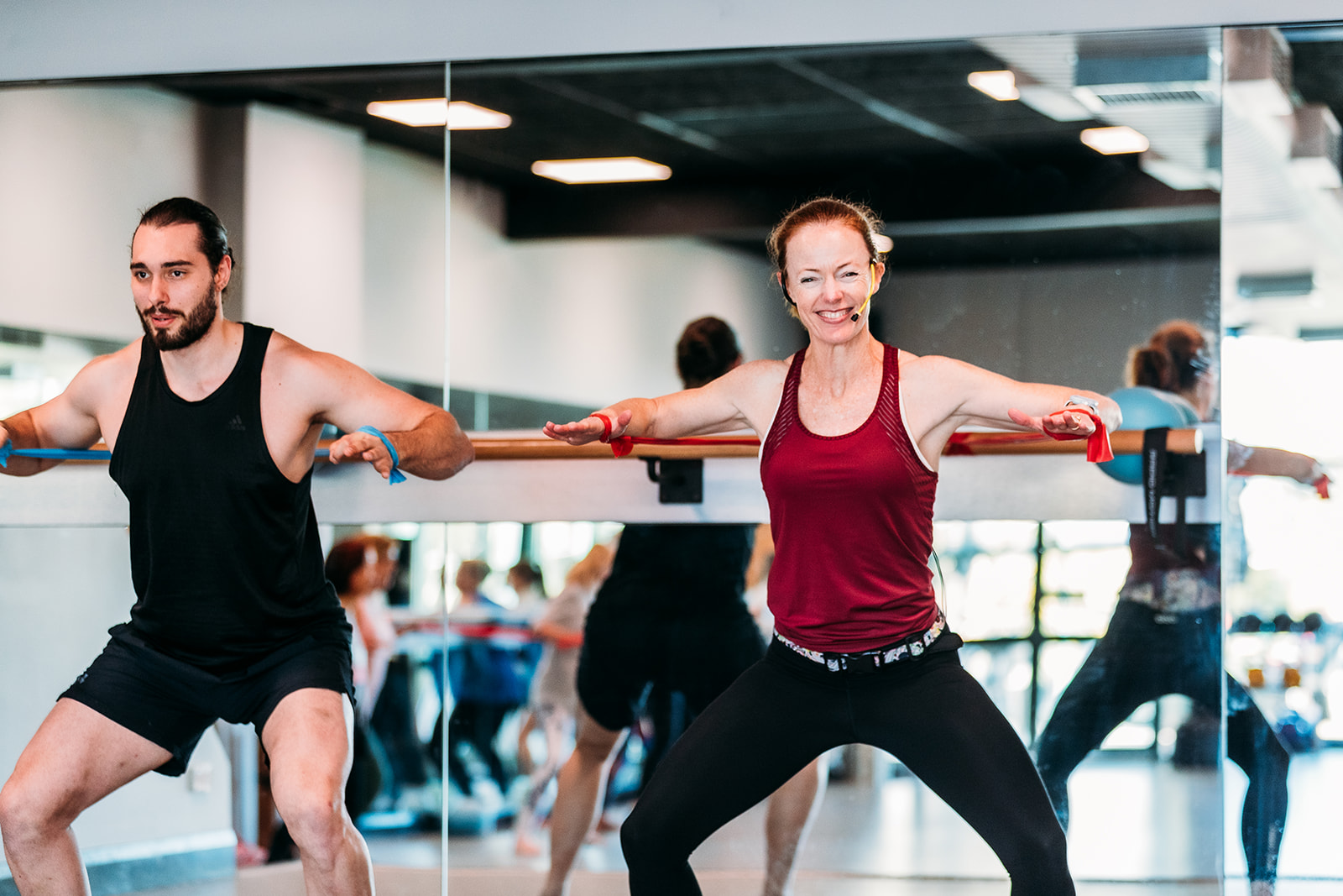 A male instructor with a beard in a black singlet next to a woman in a red singlet both in a squat in front of a ballet bar and mirror