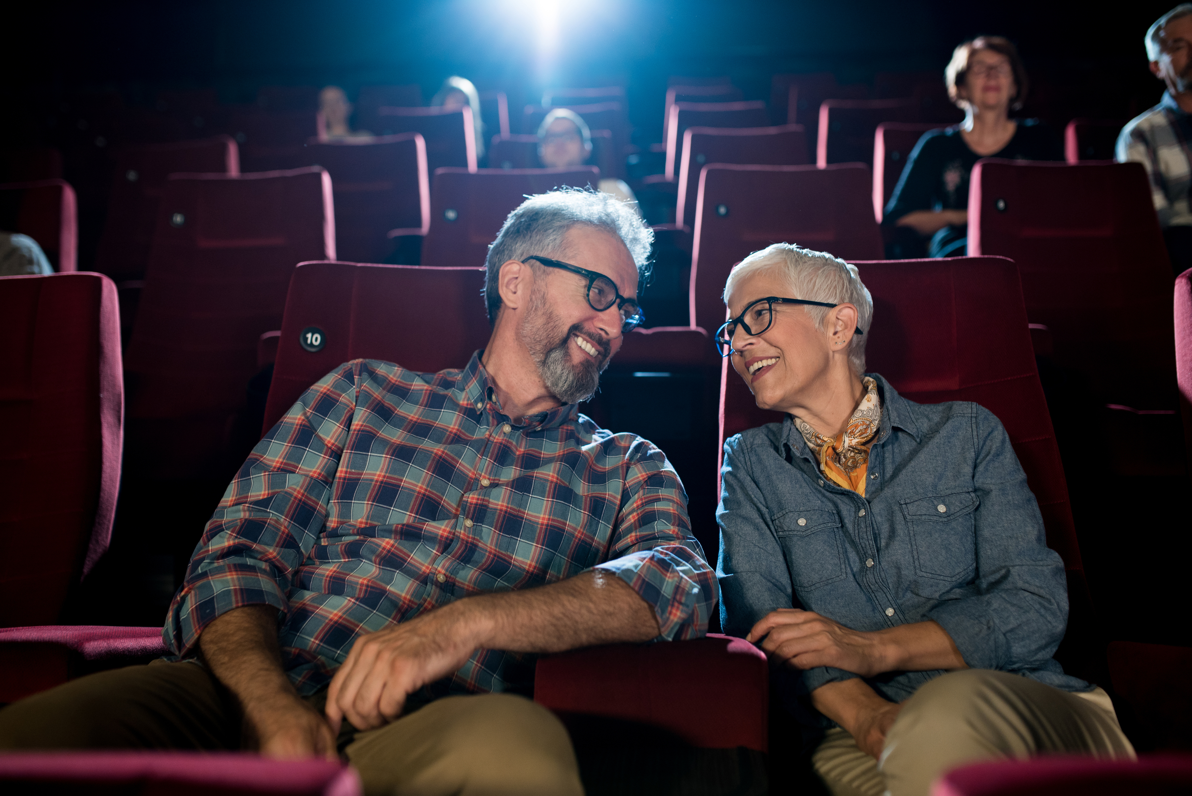 A couple in their sixties sitting on red cinema seats, they are leaning towards each other smiling.
