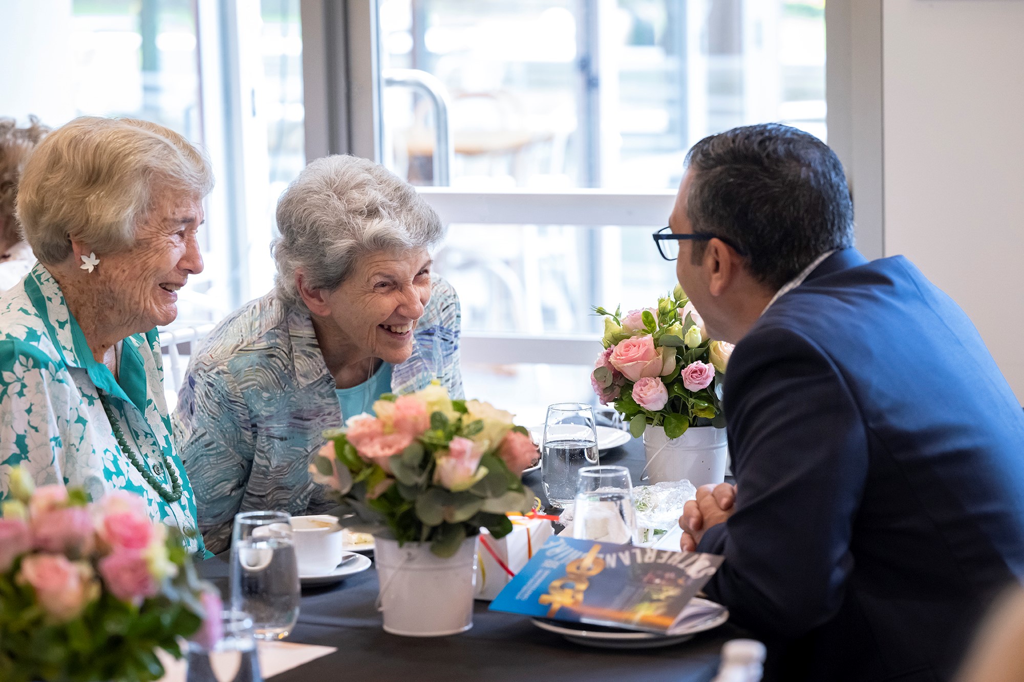 Two delighted elderly ladies having morning tea talking to the Mayor.  The table is set with lovely bouquets of flowers. 